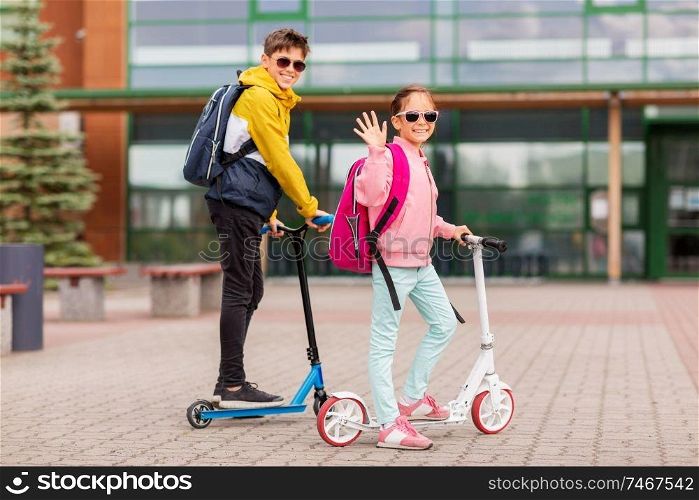 education, childhood and people concept - happy school children with backpacks riding scooters outdoors. school children with backpacks riding scooters