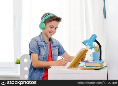 education, childhood and people concept - happy student boy in headphones writing to notebook from book at home. student boy in headphones writing to notebook