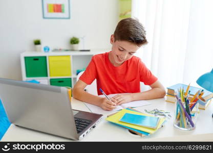 education, childhood and people concept - happy student boy with laptop computer writing to notebook at home. student boy with laptop writing to notebook