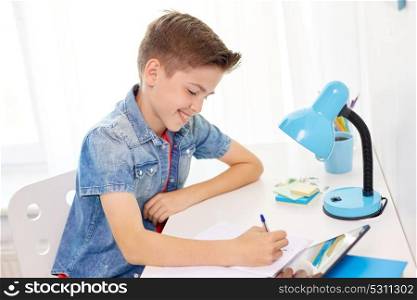 education, childhood and people concept - happy student boy with tablet pc writing to notebook at home. happy student boy writing to notebook at home