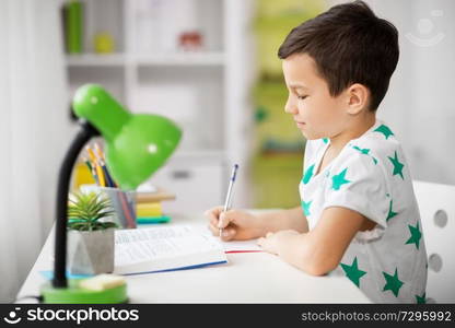 education, childhood and school concept - boy with book writing to notebook at home. boy with book writing to notebook at home