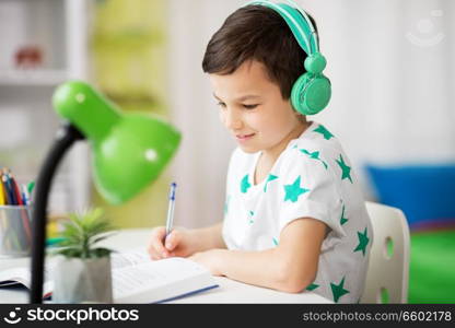 education, childhood and school concept - happy student boy in headphones writing to notebook from book at home. boy in headphones with textbook learning at home