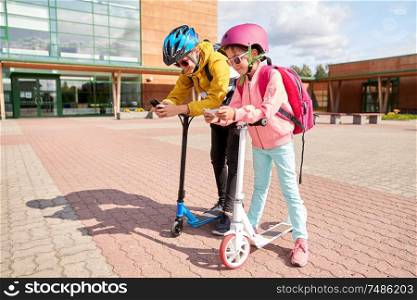education, childhood and technology concept - happy school children in helmets with smartphones and scooters outdoors. school children with smartphones and scooters