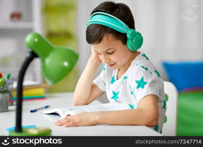education, childhood and technology concept - happy schoolboy in headphones reading book at home. boy in headphones with textbook learning at home