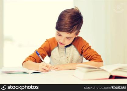 education, childhood, people, homework and school concept - smiling student boy with book writing to notebook at home. smiling student boy writing to notebook at home