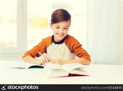 education, childhood, people, homework and school concept - smiling student boy with book writing to notebook at home. smiling student boy writing to notebook at home