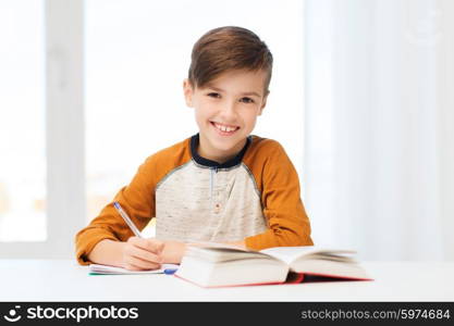 education, childhood, people, homework and school concept - smiling student boy with book writing to notebook at home