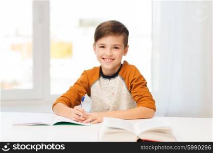 education, childhood, people, homework and school concept - smiling student boy with book writing to notebook at home