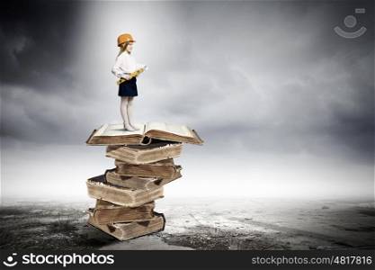 Education concept. Image of cute school girl in hardhat standing on pile of books