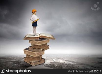 Education concept. Image of cute school girl in hardhat standing on pile of books