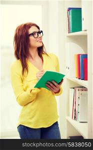 education concept - smiling redhead female student in eyeglasses with textbook in library
