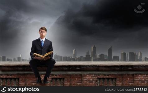 Education concept. Young businessman sitting on building top with book