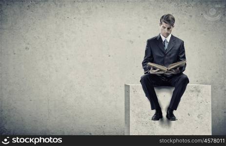 Education concept. Young businessman sitting on granite cube with book