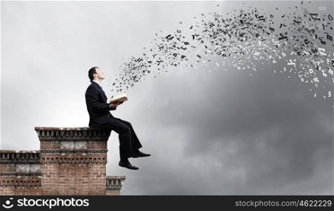 Education concept. Young businessman sitting on top of building and reading book