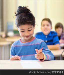 education, elementary school and children concept - happy little student girl with pen and paper writing over classroom and classmates background