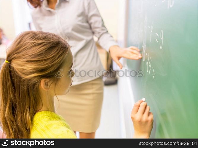 education, elementary school, learning, math and people concept - close up of little schoolgirl with teacher writing on chalk board and solving task at classroom