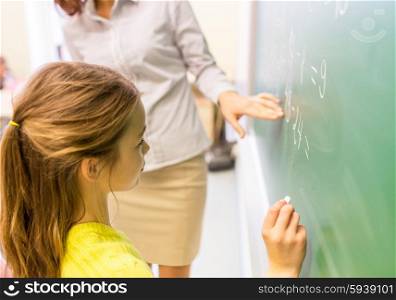 education, elementary school, learning, math and people concept - close up of little schoolgirl with teacher writing on chalk board and solving task at classroom