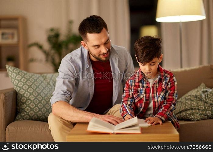 education, family and homework concept - happy father and son with book writing to notebook at home in evening. father and son doing homework together