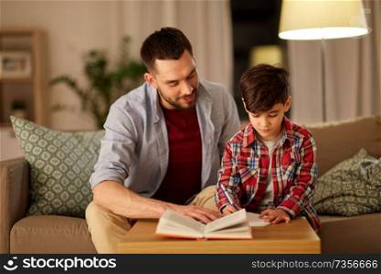 education, family and homework concept - happy father and son with book writing to notebook at home in evening. father and son doing homework together