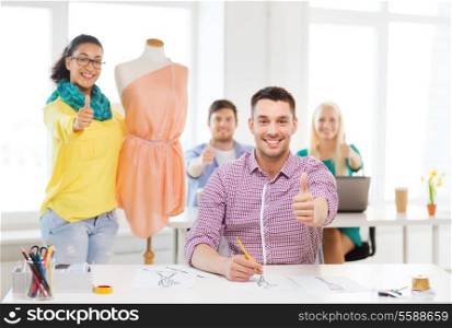 education, fashion and office concept - smiling male drawing sketches and female adjusting dress on mannequin in office and showing thumbsup