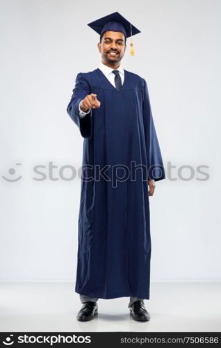 education, graduation and choice concept - happy smiling indian male graduate student in mortar board and bachelor gown pointing to you over grey background. indian graduate student pointing to you