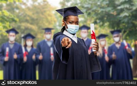 education, graduation and pandemic concept - graduate student woman in mortarboard and bachelor gown with diploma wearing protective mask pointing to camera over group of people on background. female graduate student in mask with diploma