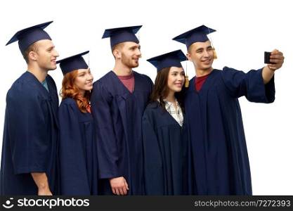education, graduation and people concept - group of happy graduate students in mortar boards and bachelor gowns taking selfie by smartphone over white background. graduates taking selfie by smartphone