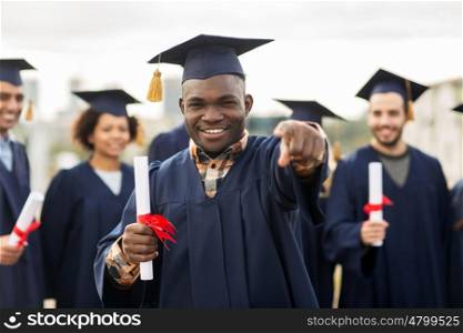 education, graduation and people concept - group of happy international students in mortar boards and bachelor gowns with diplomas pointing finger at you