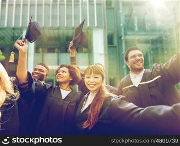 education, graduation and people concept - group of happy international students in bachelor gowns waving mortar boards or hats over university building background