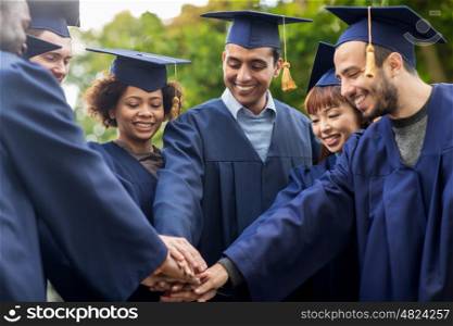 education, graduation and people concept - group of happy international students in mortar boards and bachelor gowns putting hands together