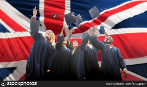 education, graduation and people concept - group of happy international students in bachelor gowns throwing mortarboards up over english flag background