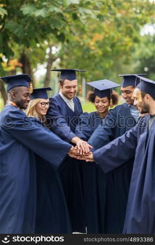 education, graduation and people concept - group of happy international students in mortar boards and bachelor gowns putting hands on top