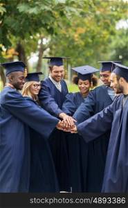 education, graduation and people concept - group of happy international students in mortar boards and bachelor gowns putting hands on top
