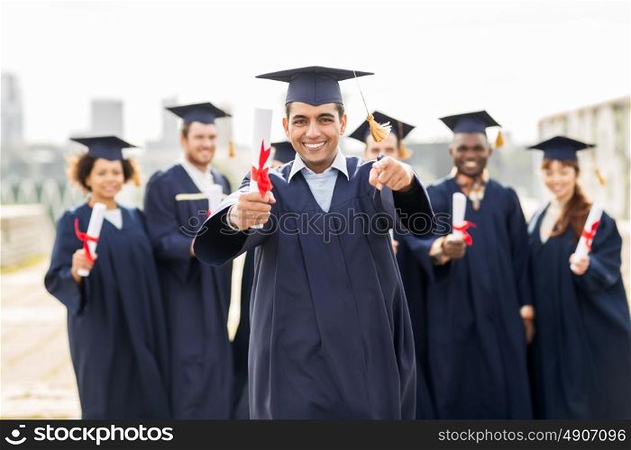 education, graduation and people concept - group of happy international students in mortar boards and bachelor gowns with diplomas pointing finger at you. happy student with diploma pointing finger at you