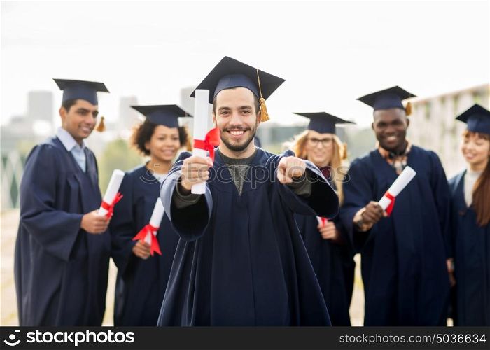 education, graduation and people concept - group of happy international students in mortar boards and bachelor gowns with diplomas pointing finger at you. happy student with diploma pointing finger at you