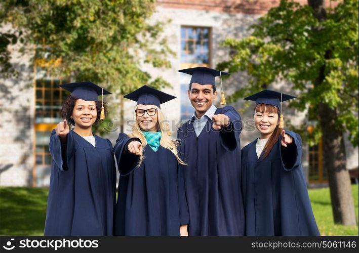 education, graduation and people concept - group of happy international students in mortar boards and bachelor gowns pointing finger at you over campus building background. happy students or bachelors pointing finger at you