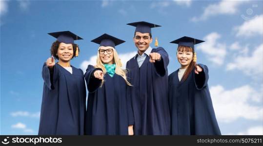 education, graduation and people concept - group of happy international students in mortar boards and bachelor gowns pointing finger at you over blue sky and clouds background. happy students or bachelors pointing finger at you