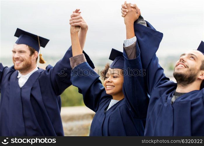 education, graduation and people concept - group of happy international students in mortar boards and bachelor gowns holding hands and celebrating success. happy students celebrating graduation