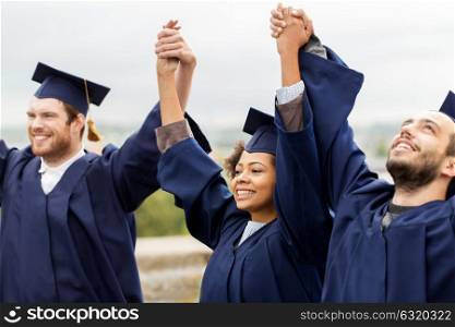 education, graduation and people concept - group of happy international students in mortar boards and bachelor gowns holding hands and celebrating success. happy students celebrating graduation