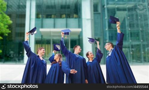 education, graduation and people concept - group of smiling students in gowns waving mortarboards over school or university building background