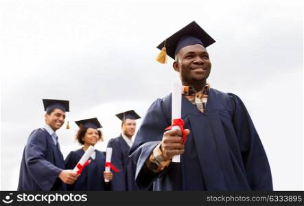 education, graduation and people concept - happy african american graduate in mortar boards and bachelor gown with diploma and group of international students. happy students in mortar boards with diplomas