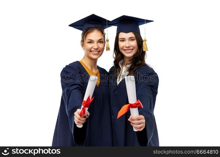 education, graduation and people concept - happy female graduate students in mortar boards and bachelor gowns with diplomas over white background. female graduates in mortar boards with diplomas