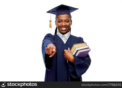 education, graduation and people concept - happy graduate student woman in mortarboard and bachelor gown with books pointing finger to camera over white background. happy female graduate student with books