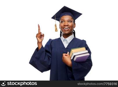 education, graduation and people concept - happy graduate student woman in mortarboard and bachelor gown with books pointing finger up over white background. happy female graduate student with books