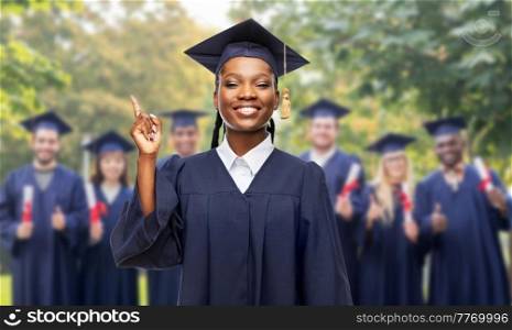 education, graduation and people concept - happy graduate student woman in mortarboard and bachelor gown pointing finger up over group of bachelors at park on background. happy female graduate student pointing finger up