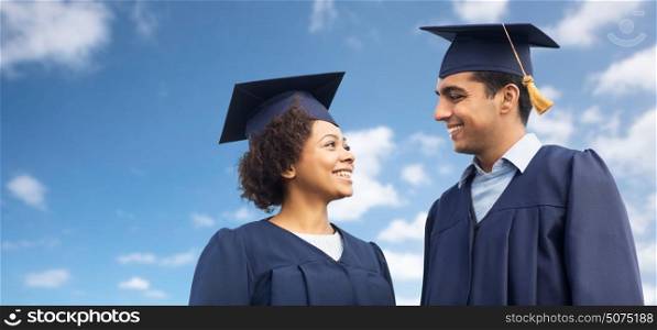education, graduation and people concept - happy international students in mortar boards and bachelor gowns over blue sky and clouds background. happy students or bachelors in mortar boards