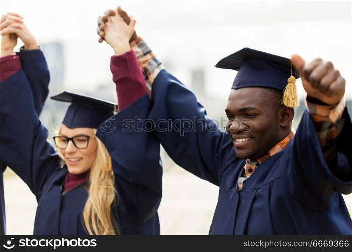 education, graduation and people concept - happy international students in mortar boards and bachelor gowns holding hands and celebrating success. happy students celebrating graduation. happy students celebrating graduation