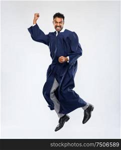 education, graduation and people concept - happy smiling indian male graduate student in bachelor gown celebrating success and jumping over grey background. happy indian graduate student celebrating success