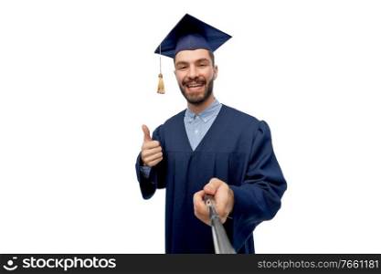 education, graduation and people concept - happy smiling male graduate student in mortar board and bachelor gown taking picture with selfie stick over white background. male graduate student taking selfie with monopod
