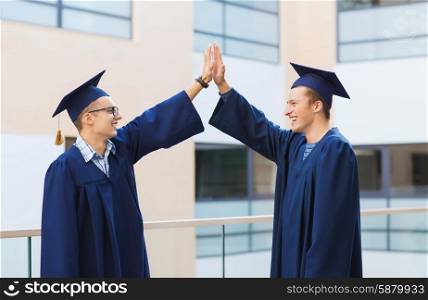 education, graduation and people concept - smiling students in mortarboards and gowns making high five gesture outdoors
